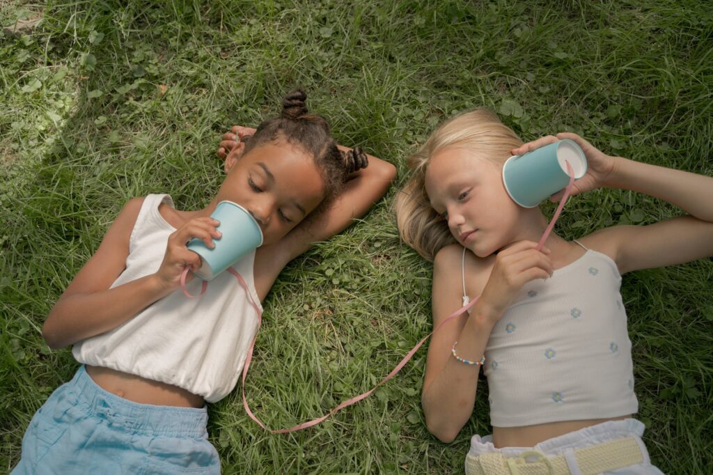 two teenage girls laying on grass and playing telephone call using paper cups on string